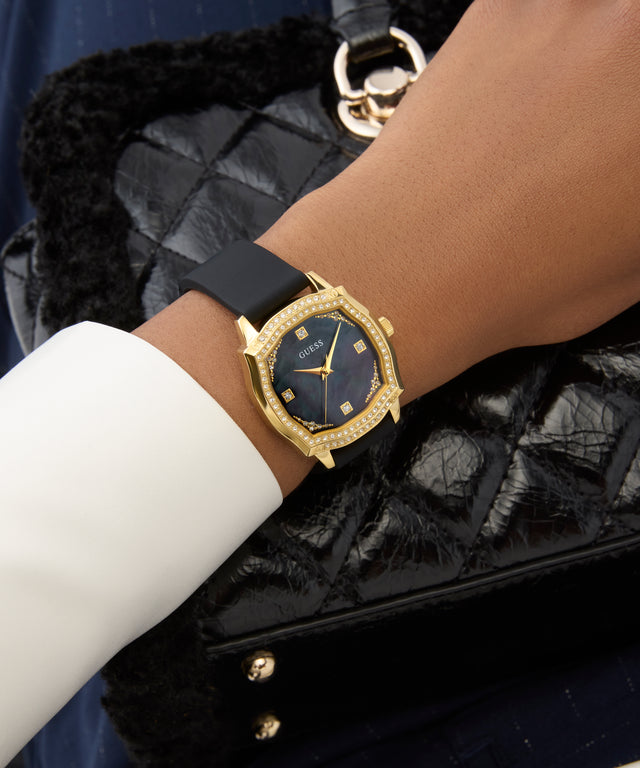 GUESS Ladies Black Gold Tone Analog Watchon a wrist with a black strap against a black quilted handbag background