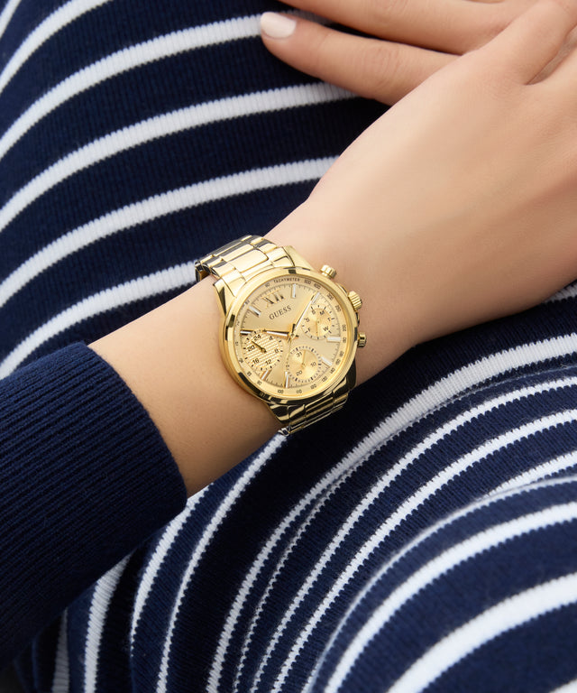 Gold GUESS Ladies Gold Tone Multi-function Watch on a person's wrist with a striped background