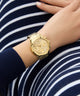 Gold GUESS Ladies Gold Tone Multi-function Watch on a person's wrist with a striped background