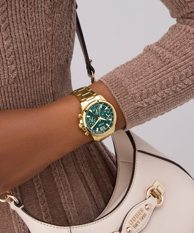 Gold GUESS Ladies Gold Tone Multi-function Watch with green face on wrist, wearing a brown sweater, holding a white handbag.