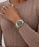 Person wearing a gold and green GUESS Ladies 2-Tone Multi-function Watch on a brown sweater
