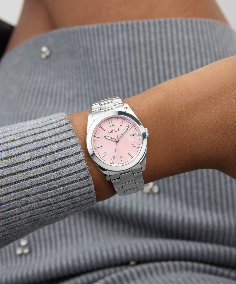 Wrist wearing a GUESS Ladies Silver Tone Date Watch with a pink face on a gray textured sleeve.
