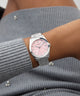 Wrist wearing a GUESS Ladies Silver Tone Date Watch with a pink face on a gray textured sleeve.