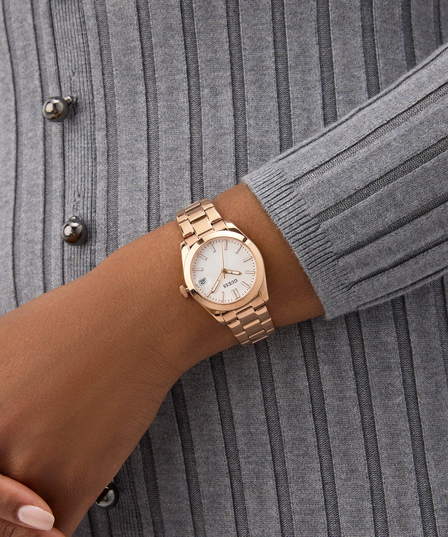 Rose gold GUESS Ladies Rose Gold Tone Date Watch on a wrist with a gray textured background