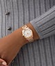 Rose gold GUESS Ladies Rose Gold Tone Date Watch on a wrist with a gray textured background