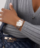 Person wearing a gold GUESS Ladies 2-Tone Date Watch with a gray sweater and Guess handbag