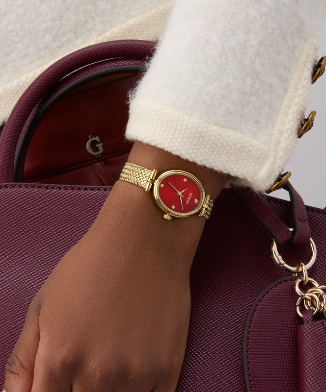 Gold watch with a red face on a wrist, wearing a white coat, with a maroon handbag in the background.