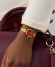 Gold watch with a red face on a wrist, wearing a white coat, with a maroon handbag in the background.