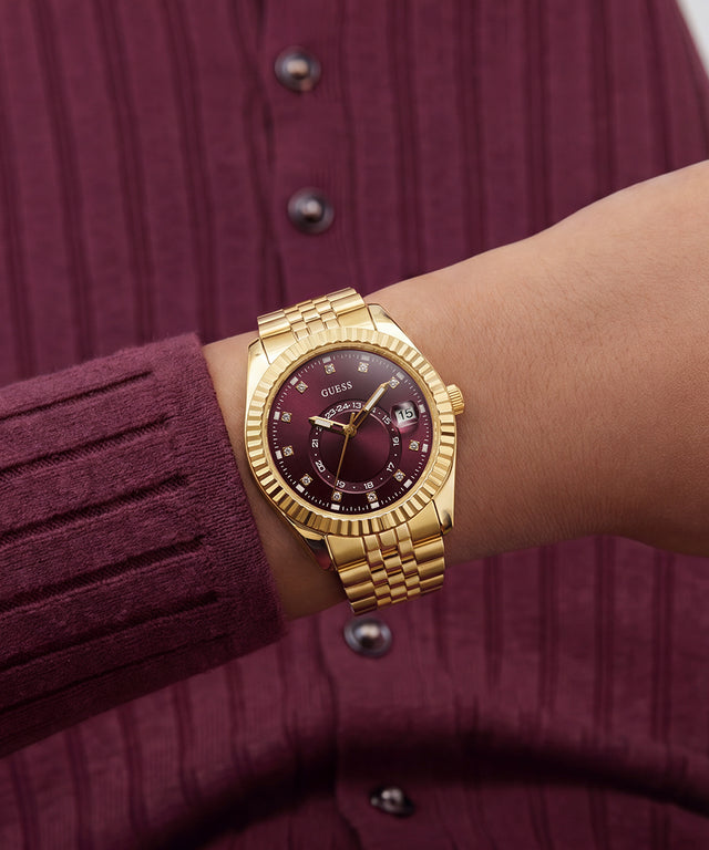 A gold-tone wristwatch with a dark burgundy dial and date window, worn on a person's wrist over a ribbed burgundy cardigan.