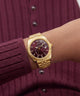 A gold-tone wristwatch with a dark burgundy dial and date window, worn on a person's wrist over a ribbed burgundy cardigan.