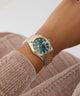 Gold and silver watch with a green face on a wrist, wearing a beige sweater. GW0936L3