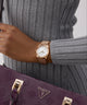 Person wearing a GUESS Ladies Rose Gold Tone Multi-function Date Watchwith a gray sweater and purple handbag featuring a brand logo.