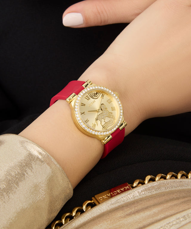 Gold GUESS Ladies Limited Edition Lunar New Year Red Gold Tone Analog Watch with a red strap on a wrist, with a gold clutch in the background.