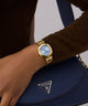Gold GUESS Ladies Brown Gold Tone Analog Watch on a wrist with a blue handbag featuring a brand logo.