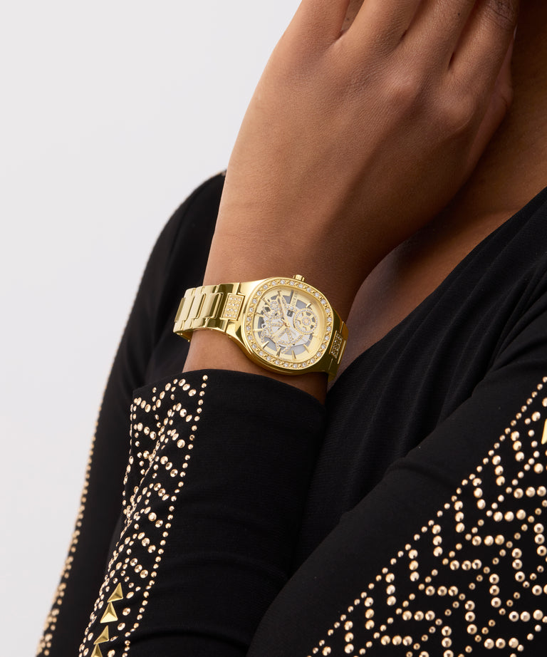 GUESS Ladies Gold Tone Analog Watch on a person's wrist with a black garment featuring gold patterns.
