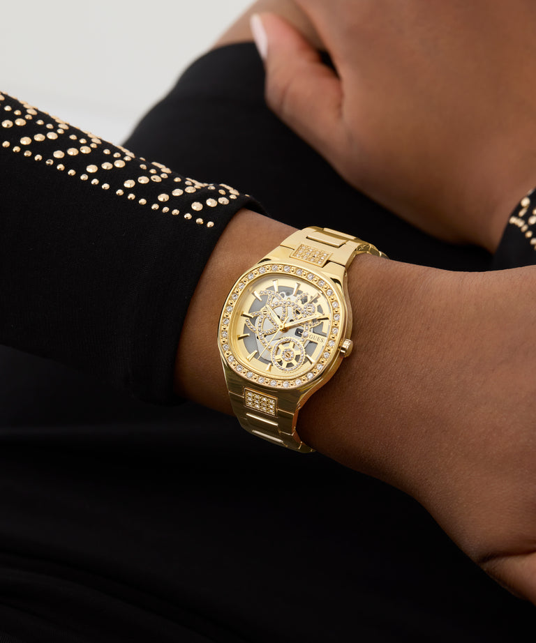 GUESS Ladies Gold Tone Analog Watch with diamond accents on a person's wrist against a neutral background