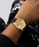 GUESS Ladies Black Gold Tone Analog Watch with a triangle design on a wrist against a black sequined background