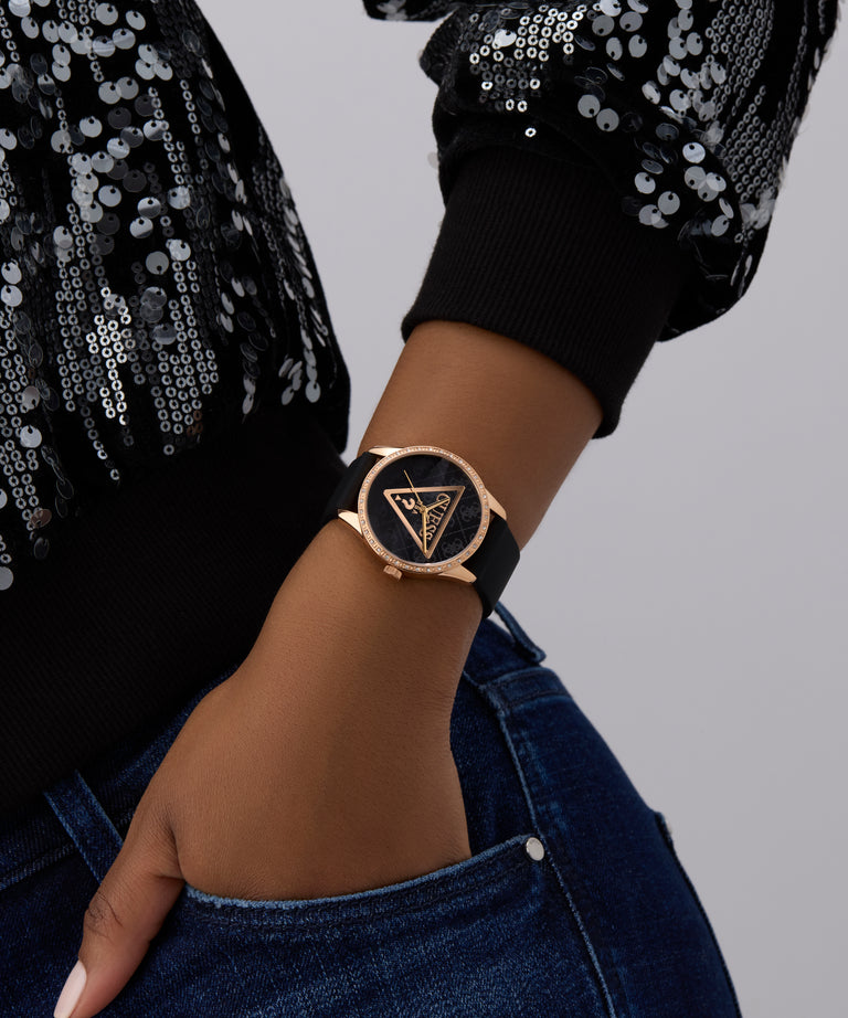 Person wearing a black GUESS Ladies Black Rose Gold Tone Analog Watch with a gold triangle design on a gray background