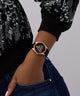 Person wearing a black GUESS Ladies Black Rose Gold Tone Analog Watch with a gold triangle design on a gray background