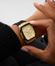 Gold watch with black strap on a wrist against a neutral background