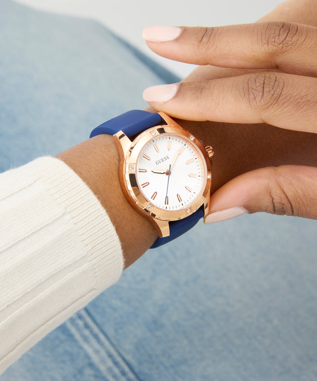 Hand wearing a GUESS Ladies Blue Rose Gold Tone Analog Watch with a blue strap and gold case, against a blurred background.