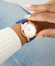 Hand wearing a GUESS Ladies Blue Rose Gold Tone Analog Watch with a blue strap and gold case, against a blurred background.