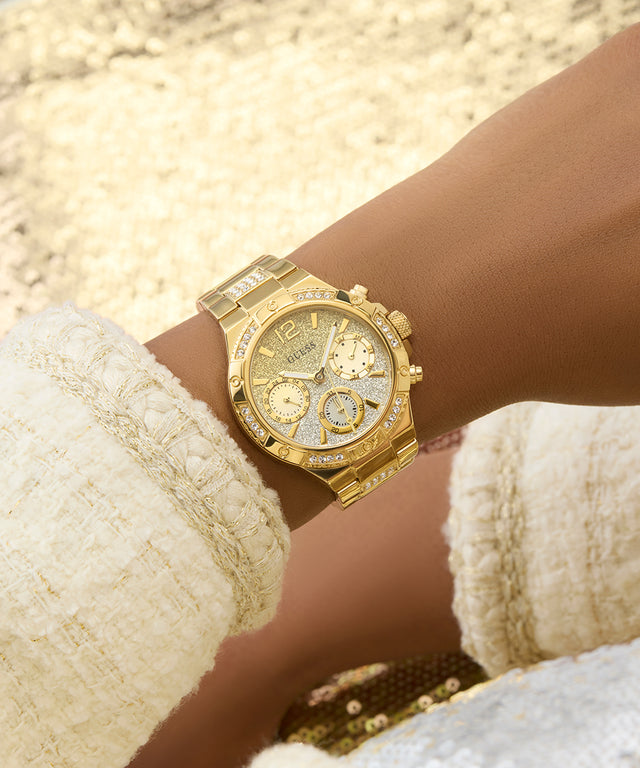 GUESS Ladies Gold Tone Multi-function Watch with diamond accents on a wrist, wearing a white textured garment.