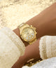 GUESS Ladies Gold Tone Multi-function Watch with diamond accents on a wrist, wearing a white textured garment.