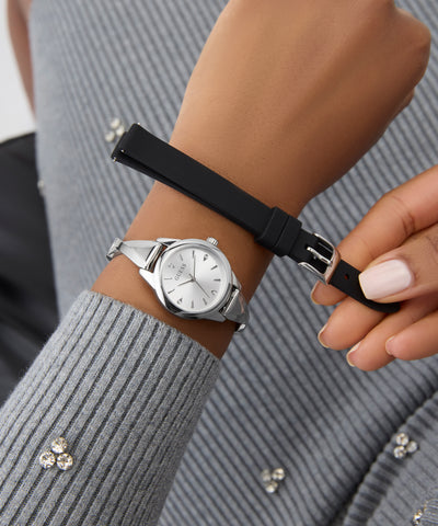 Person wearing a GUESS Ladies Silver Tone Analog Watch with a black strap on a gray textured sleeve.