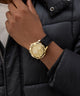 Gold GUESS Mens Black Gold Tone Multi-function Watch with a black strap on a person wearing a black puffer jacket.