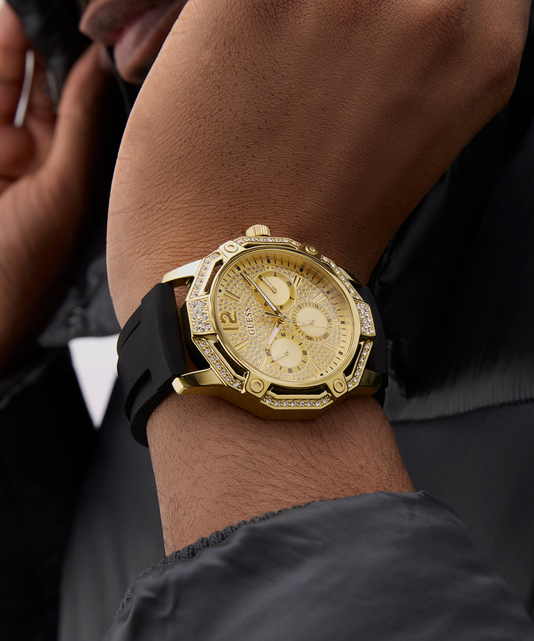 Gold GUESS Mens Black Gold Tone Multi-function Watch with black strap on a wrist against a dark background