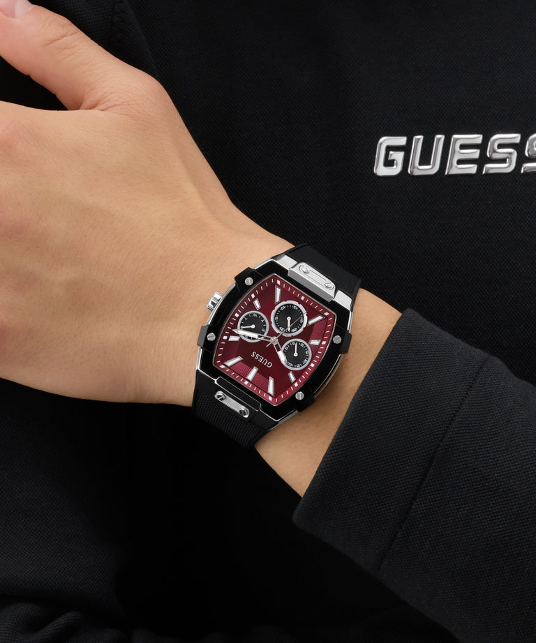 Hand wearing a black and red GUESS Mens Black Silver Tone Analog Watch with a Guess logo in the background