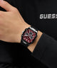 Hand wearing a black and red GUESS Mens Black Silver Tone Analog Watch with a Guess logo in the background