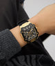 Gold and black watch on a wrist with a blurred background GUESS Mens Black Gold Tone Multi-function Watch GW0956G2