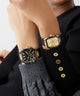 Two gold and black watches on a person's wrist against a neutral GUESS Mens Black Gold Tone Multi-function Watch GW0956G2background