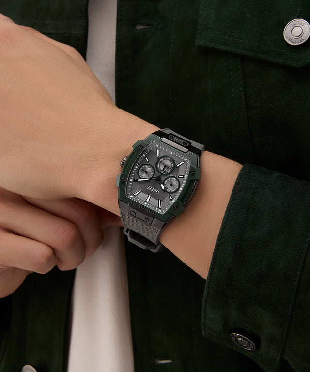 Person wearing a green watch with a detailed face on a wrist, against a dark jacket background. GUESS Mens Grey Gunmetal Multi-function Watch