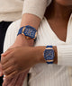 Two wrists wearing blue and gold watches on a neutral background