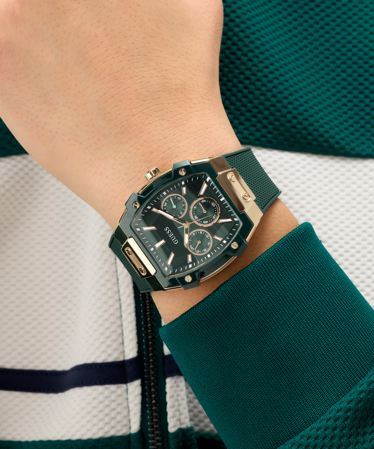 Close-up of a person's wrist wearing a black and gold GUESS Mens Green Rose Gold Tone Multifunction Watch with a green strap.