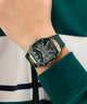 Close-up of a person's wrist wearing a black and gold GUESS Mens Green Rose Gold Tone Multifunction Watch with a green strap.