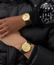 Person wearing a gold GUESS Mens Black Gold Tone Multi-function Watch with a black strap on a black background