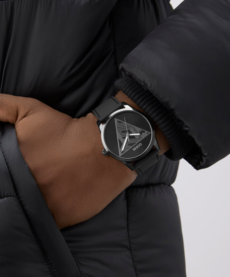 Person wearing a black puffer jacket with a black GUESS Mens Black Analog Watch on a neutral background