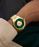 GUESS Mens Gold Tone Analog Watch with green face on a wrist, wearing a black shirt and beige jacket.