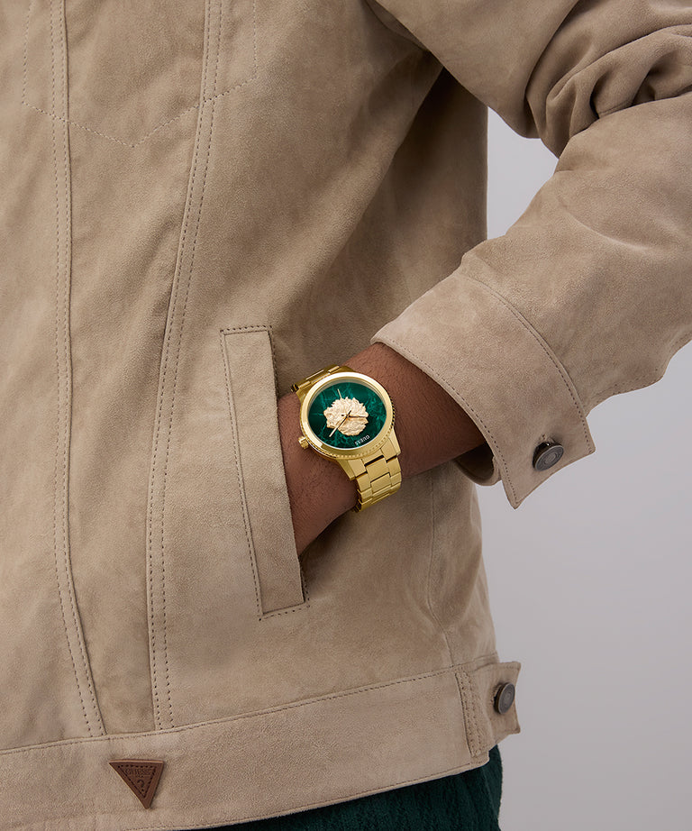 Close-up of a person wearing a gold GUESS Mens Gold Tone Analog Watch with a green face on a beige jacket.