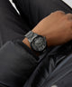 Close-up of a person wearing a dark gray watch with intricate design on a black background