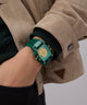 Person wearing a GUESS Mens Green Analog Watch with decorative elements on a plain background