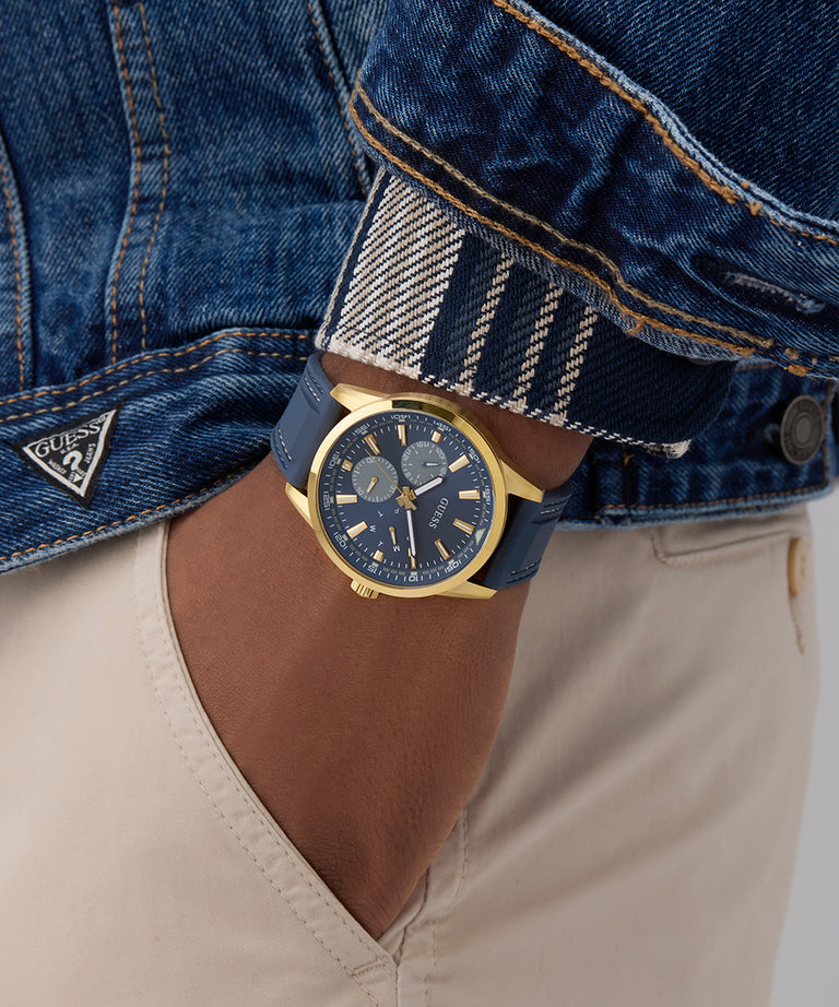 Blue watch with gold accents on a person wearing a denim jacket and beige pants. GUESS Mens Blue Gold Tone Multi-function Watch GW0964G2