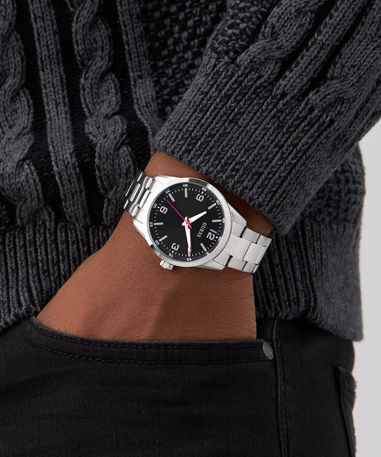 Hand wearing a GUESS Mens Silver Tone Analog Watch with a black face, against a gray sweater background