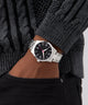 Hand wearing a GUESS Mens Silver Tone Analog Watch with a black face, against a gray sweater background