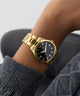 Gold wristwatch with a dark face on a person's wrist against a neutral background GW0965G2