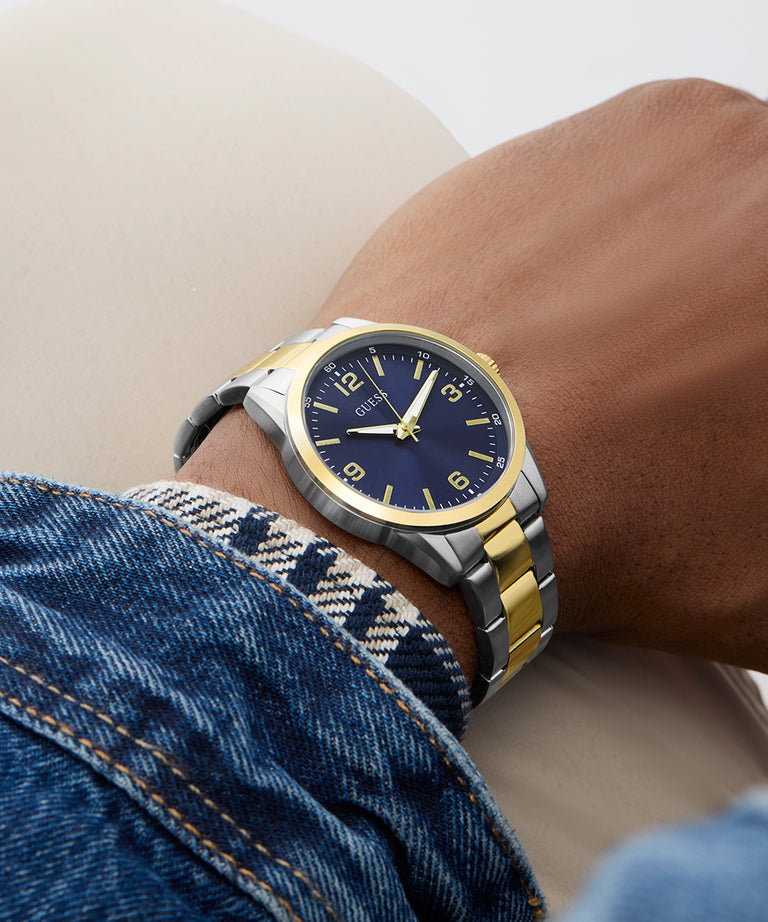 Wrist wearing a watch with a blue face and gold accents, with denim jeans in the background. GW0965G4